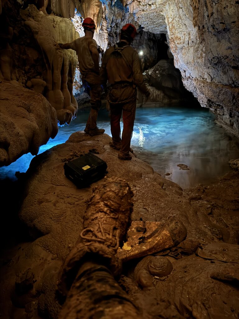 Muddy boot in the foreground of photo while two cavers look at a pool lit with a bluish light.